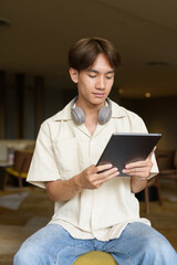 Non binary Asian student man sitting in modern coffee shop using digital tablet computer