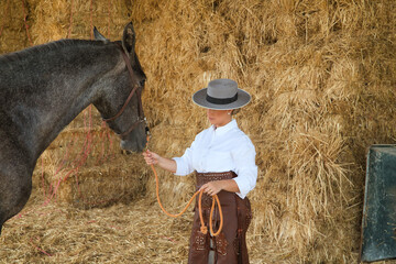 Young, beautiful, blonde woman in white shirt, leather chaps and hat, holding and watching her horse, with hay bales in the background. Concept animals, riding, horses, sport.