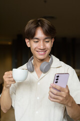 Non binary Asian student man sitting in cafe drinking coffee and using phone