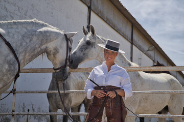 Young, beautiful, blonde woman in white shirt, leather chaps, hat and a riding crop in her hands, posing looking at camera in a stable surrounded by horses. Concept animals, riding, horses, sport.