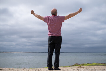 Elderly man breathing deeply with arms outstretched by the sea, back view