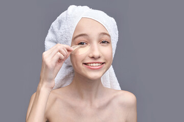 Young caucasian female applying under eye patch with towel on head against gray background.