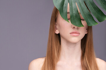 Young caucasian female with long hair and closed eyes partially covered by leaf on grey background.