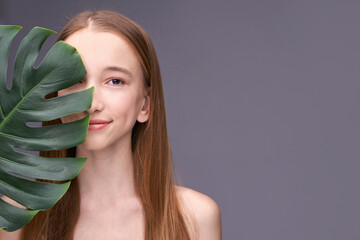 Young caucasian female with long hair holding leaf in minimalist portrait on gray background.