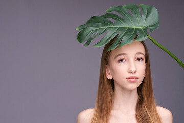 Young caucasian teen girl with long hair posing behind green monstera leaf on gray background.