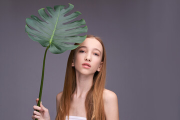 Young caucasian teen female holding large leaf against gray background with long straight hair and natural expression.