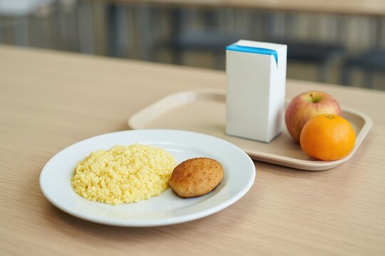 School cafeteria scene showing a freshly made lunch with millet porridge, a chicken cutlet on a plate, alongside a milk carton and fruits on a tray, with a blank space for text.
