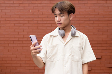 Non binary cute Asian young gay man using mobile phone against brick wall