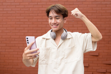 Non binary cute Asian young gay man using mobile phone against brick wall