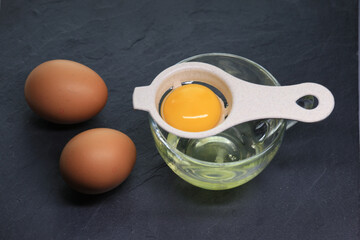 A shot of a beige egg separator holding a bright yellow yolk above egg whites in a clear glass cup, with two fresh whole eggs on a dark black surface.