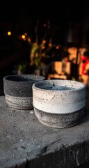 Two handmade, speckled, grey and white ceramic plant pots rest on a wooden surface.  Blurred background suggests a market or indoor setting with warm lights and greenery