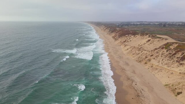 Drone flies laterally over ocean waves, sandy beach, and dunes at Fort Ord Dunes State Park, Marina, California.