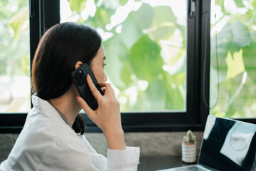 Fototapeta premium Professional woman multitasking with phone and laptop in a bright office setting, surrounded by natural greenery.