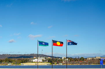 Torres Strait Islander flag, Aboriginal flag, and Australian national flag in front of Lake Burley Griffin with Parliament House in the background in Canberra, Australia.
