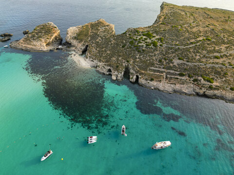Aerial view of turquoise waters meeting the rugged coastline, where boats gently float near the sandy beach, Comino, Gajnsielem, Malta.