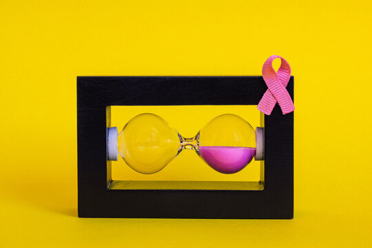 Creative visual of a horizontal hourglass filled with pink sand and a pink ribbon attached to the frame, symbolizing breast cancer awareness on a vibrant yellow background, represents paused time 