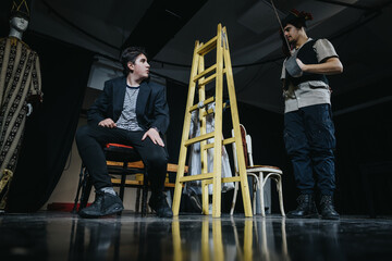Two young actors perform on a theater stage, surrounded by props and costumes, showcasing...