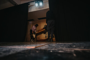 Stage crew members organizing props behind curtains in a theater setting. Light shines on the dark...