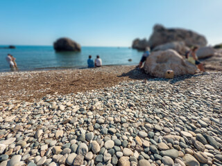 Smooth pebble beach with natural stones and people relaxing by the sea on a sunny day, showing textures of round rocks, hand holding stone and coastal landscape © Katarzyna Ledwoń