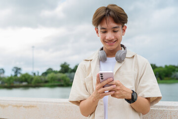 Asian non binary tourist person with headphones relaxing at park near lake using mobile phone