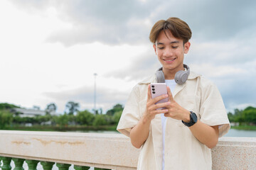 Asian non binary tourist person with headphones relaxing at park near lake using mobile phone
