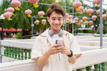 Asian non binary tourist person with headphones relaxing at park near lake using mobile phone