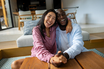 Excited child-free multiethnic couple embracing and laughing in modern living room, calm lifestyle