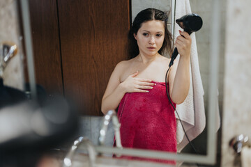 A young woman standing in front of a bathroom mirror, wearing a towel and blow-drying her hair,...