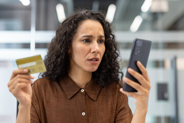 A woman looks concerned while holding a credit card and a smartphone, possibly experiencing...