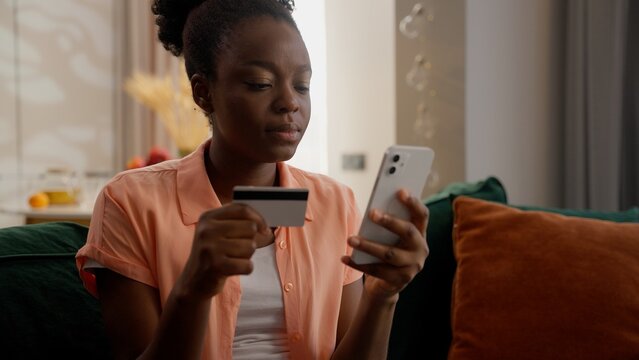 Confused upset African American ethnic business woman female girl businesswoman homeowner holding credit card using mobile phone fail online pay bank app error no money scam fraud transaction at home