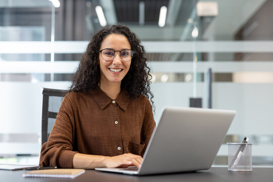 A smiling woman with curly hair and glasses works on a laptop at her desk in an office setting.
