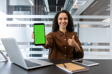 A woman in a modern office setting holds up a phone with a green screen, giving a thumbs-up gesture.