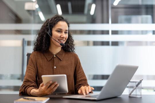 A smiling woman with curly hair wearing a headset uses a tablet and laptop while working at a desk in an office setting.
