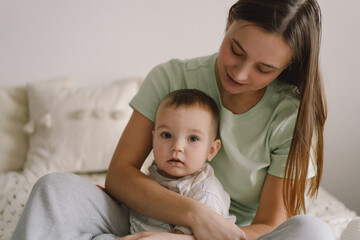 Home portrait of a baby boy with mother on the bed relaxing and playing in the bed at the weekend together, lazy morning, warm and cozy scene. Mom holding and kissing her child. Mother day concept