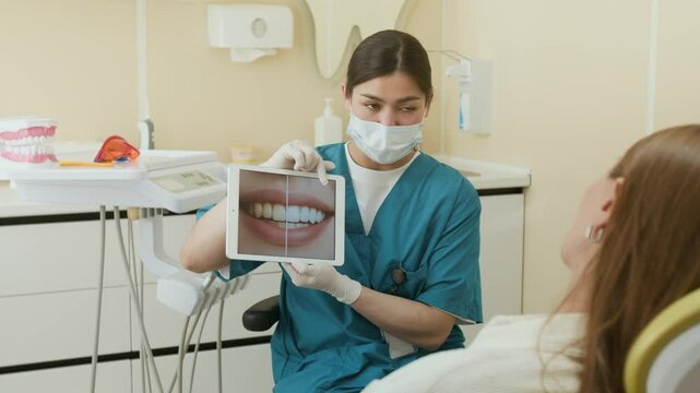 Medium shot of young female dentist holding digital tablet and explaining teeth whitening to patient in dental clinic