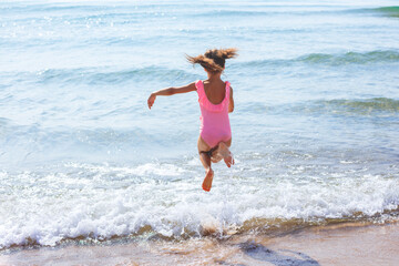 Summer on the beach. Happy girl jumping into the sea. Baby in a pink swimsuit