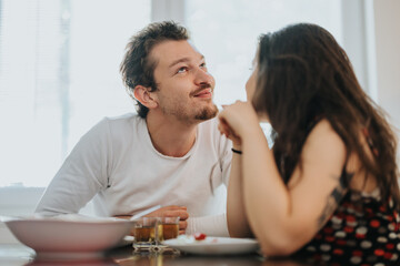 A young couple shares a tender moment at breakfast, sitting close and exchanging loving glances. The cozy setting enhances a sense of warmth and connection, highlighting their intimate bond.