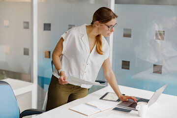 A focused woman deeply analyzing important documents at a chic office desk equipped with modern...