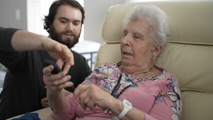 In a cozy living room, a grandson is guiding his grandmother to share photos on her phone, creating happy memories together. Their expressions show joy and connection.