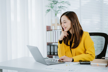 Portrait of a cheerful Asian businesswoman using a laptop and writing on a charger to keep notes as...