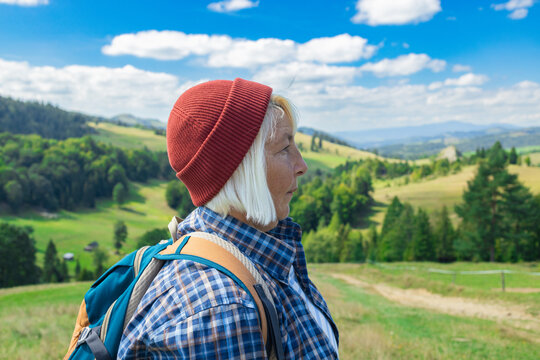Elderly woman standing on a lush green hillside with a backpack, looking content and inspired by the beautiful panoramic mountain view. Dressed in a plaid shirt and red beanie, she represents active