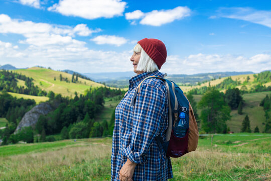Elderly woman standing on a lush green hillside with a backpack, looking content and inspired by the beautiful panoramic mountain view. Dressed in a plaid shirt and red beanie, she represents active - Powered by Adobe
