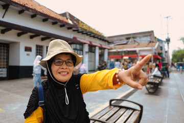 A cheerful old female tourist wearing a hat and glasses points at something in the tourist crowd of...
