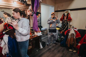 A group of individuals in a theater dressing area selecting costumes and dressing in preparation for a performance, surrounded by various garments and stage attire.