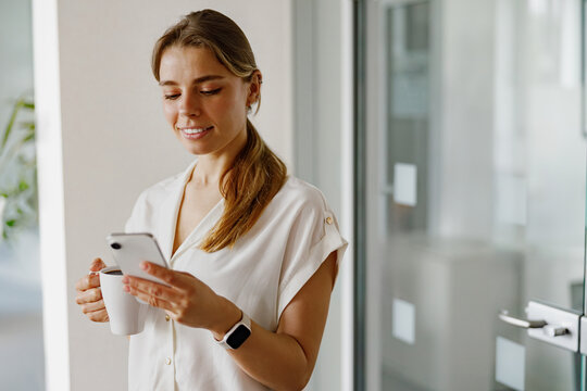 In a contemporary workspace, a woman takes pleasure in sipping her coffee while actively using her smartphone