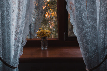 Dried flowers sit by a window with lace curtains, creating a cozy and peaceful atmosphere. A charming interior scene showcasing a vase of yellow mimosa flowers bouquet, and a view of trees outside.