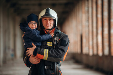 Holding kid in hands. Firefighter is with baby boy indoors