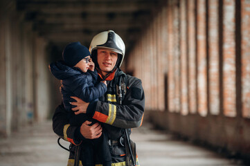 Holding kid in hands. Firefighter is with baby boy indoors