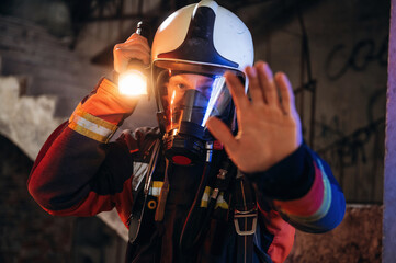 Flashlight, stop hand gesture. Firefighter in professional equipment is indoors in abandoned...