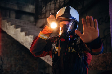 Flashlight, stop hand gesture. Firefighter in professional equipment is indoors in abandoned...